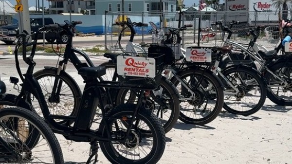 Row of rental bicycles parked on a sandy area near beach buildings.