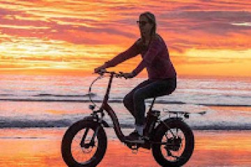 Person rides bike on beach at sunset, reflection on wet sand.