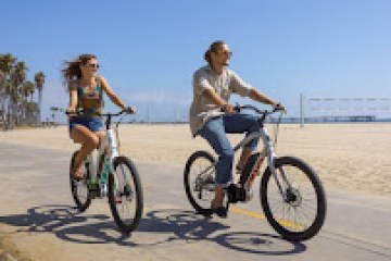Two people riding bikes on a beachside path under a clear blue sky.